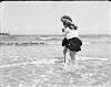 [Two girls playing in shallow water at the seaside with backs to the camera]