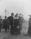 [Man and woman standing on a pavement, among other bystanders, with tram in the background]