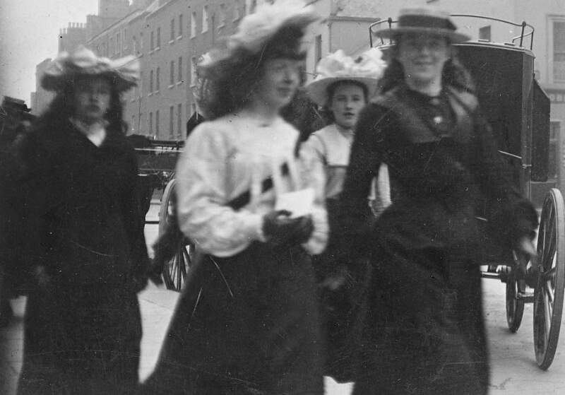 [Four women walking, with carriage in the background]