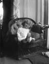 [1907. Toddler (Little girl) sitting on wicker chair, wears embroidery anglaise dress, sailor doll on seat, similar to image Clonbrock 1299.]